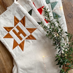 May include: Three off-white fabric stockings featuring quilted star patterns in burnt orange, bright red, and sage green. Each stocking includes a hanging loop at the top and is decorated with a thin string of small, glowing warm white LED lights.