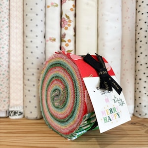 May include: A jelly roll of fabric strips featuring a variety of colourful patterns in red, green, and white. A black ribbon secures the roll, attached to a white tag with the text Ruby Society by Kimberly Kight and the words Merry Happy.