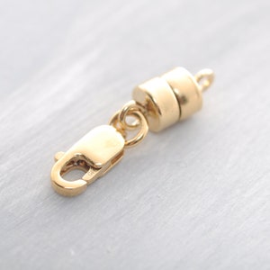 Puede incluir: Un cierre magnético dorado con un cierre de mosquetón y un cierre magnético cilíndrico. El cierre está diseñado para la fabricación de joyas y tiene pequeños bucles para sujetar a cadenas u otros componentes.