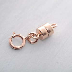 Könnte beinhalten: Ein roségoldener Magnetverschluss mit Federringverschluss. Der Verschluss hat eine zylindrische Magnetkomponente und eine kleine Schlaufe zur Befestigung an Schmuck. Das Metall hat eine glänzende Oberfläche.
