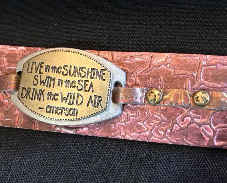 Może przedstawiać: Ręcznie robiona sk&oacute;rzana bransoletka z metalową płytką z cytatem "Live in the Sunshine, Swim in the Sea, Drink the Wild Air - Emerson." Bransoletka ma teksturowane, czerwonobrązowe wykończenie i mosiężne nity.