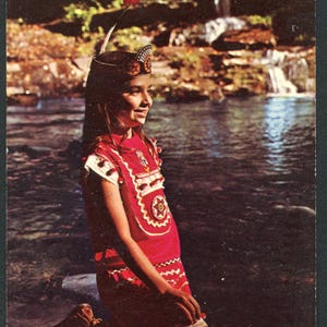 May include: A vintage postcard showing a person kneeling by a river, wearing a red Native American-style dress with white details and a decorative headpiece. The background features a waterfall and green foliage.