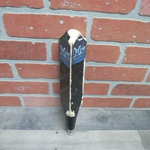 May include: A black and white tap handle with the text "D.C. O'Malley Brewing" on a blue banner. The tap handle is rectangular with a white top and a silver base. It is set against a red brick wall.