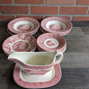 May include: A set of vintage pink and white ceramic tableware. The collection features soup bowls, saucers, and plates, all with a scenic pattern. A gravy boat with a matching rectangular base is also present. The items are displayed on a dark wooden surface.