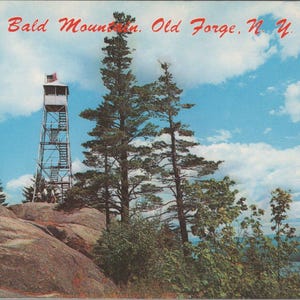 May include: Vintage postcard featuring a scenic view of Bald Mountain in Old Forge, N.Y. A tall fire tower with an American flag stands atop a rocky peak, surrounded by trees and a blue sky with clouds.