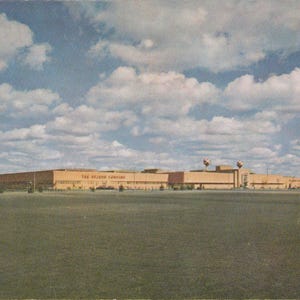 May include: A vintage postcard showing a large, tan building under a blue sky with clouds. The building is labelled "The Hecht Company". A grassy field stretches across the foreground.