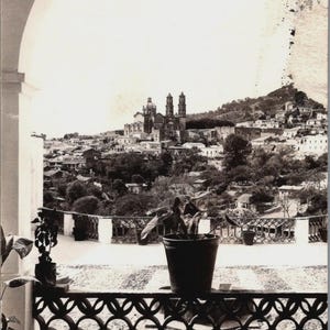 May include: Vintage black and white photograph of a cityscape, viewed from a balcony with a decorative railing. A church with twin towers dominates the hillside. A potted plant sits on the balcony. Text at the bottom reads "Taxco, Gro. 344."