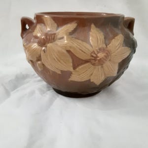 May include: A brown ceramic flower pot with two small handles. The pot features a raised floral design in shades of tan and brown. The pot is likely used for decorative purposes or for holding plants.