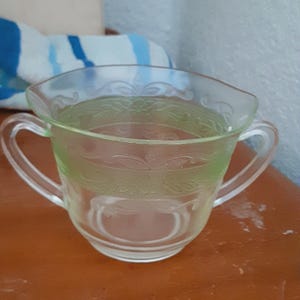 May include: A clear glass creamer with two handles. The creamer has a light green tint and a decorative etched floral pattern around the rim. The creamer is sitting on a brown surface.