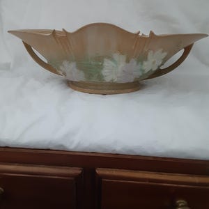 May include: A peach-colored ceramic bowl with a scalloped rim and two handles. The bowl features a floral design with white and green accents. The bowl is elevated on a circular base, suitable for decorative or serving purposes.