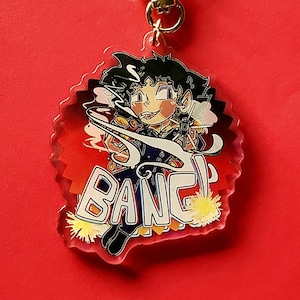 Puede incluir: Un llavero acrílico transparente con una ilustración de dibujos animados de un personaje sosteniendo un arma, con la palabra "BANG!" en blanco. El llavero tiene un cierre dorado en forma de estrella y un fondo rojo.