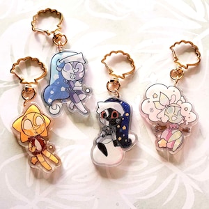 Puede incluir: Una colección de cinco llaveros acrílicos con personajes de dibujos animados. Cada llavero tiene un cierre dorado en forma de concha. Los personajes incluyen un sol, una luna y figuras con temas de nubes, todos con diseños y colores únicos.