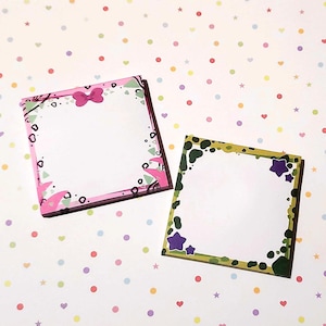 Puede incluir: Dos blocs de notas cuadrados con bordes decorativos. Uno tiene un borde rosa con un lazo, el otro un borde verde con estrellas moradas. Ambos tienen centros blancos para escribir. El fondo es blanco con lunares de colores.