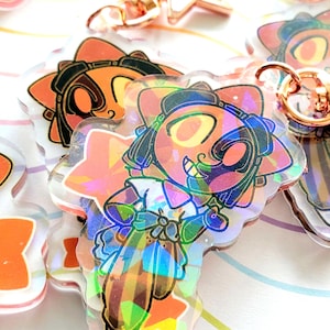 Può includere: Primo piano di diversi ciondoli olografici in acrilico raffiguranti un personaggio dei cartoni animati con capelli arancioni e una fascia nera. I ciondoli hanno una forma a fiore e sono attaccati a portachiavi color oro. Il personaggio ha un'espressione allegra.