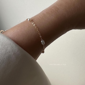May include: A gold bracelet with small, white pearls is shown on a wrist. The bracelet features a fine chain with evenly spaced pearls. The background is a neutral, off-white colour.