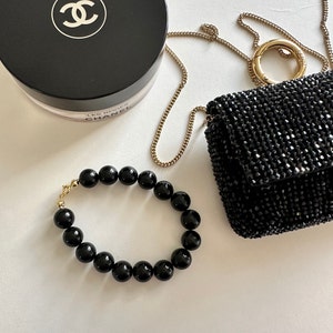 May include: A black beaded bracelet with a gold clasp. The bracelet is made of round, black beads and is lying on a white surface. There is a black purse and a Chanel compact powder in the background.