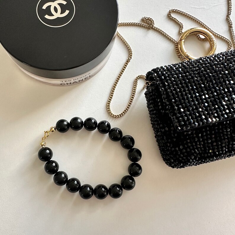 May include: A black beaded bracelet with a gold clasp. The bracelet is made of round, black beads and is lying on a white surface. There is a black purse and a Chanel compact powder in the background.