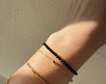 May include: A black beaded bracelet with a delicate gold chain bracelet layered on top. Both bracelets are worn on a person's wrist.
