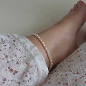 May include: A white pearl anklet on a person's ankle. The anklet is made of small, round pearls that are strung together on a thin chain. The anklet is worn on a person's ankle, which is resting on a white lace fabric.