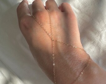 May include: A delicate gold hand chain bracelet. The jewellery piece features a fine chain that wraps around the finger and wrist, connected by a delicate chain across the back of the hand. The bracelet is displayed on a light-coloured background.