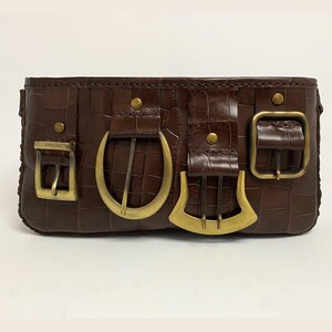 May include: A brown leather clutch with a crocodile texture. The clutch features four gold-colored buckles of varying shapes and sizes. The buckles are attached to the front of the bag with small, gold-colored rivets.