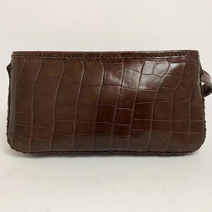 May include: A brown, rectangular clutch with a crocodile skin texture. The bag has a small handle on one side and is made of a shiny, dark brown material. The edges are stitched with a darker thread.