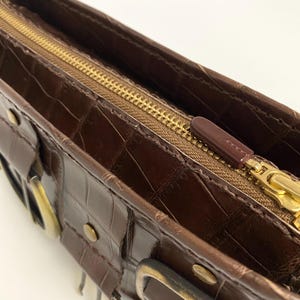 May include: A brown leather handbag with a crocodile texture. The bag features a gold zipper and hardware, including buckles and rivets. The interior is visible, showcasing the open design.
