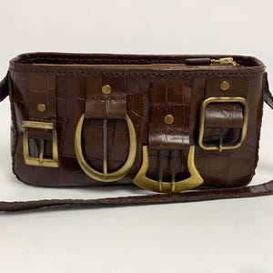 May include: A brown, rectangular handbag with a textured, crocodile-like pattern. The bag features a long, braided strap and multiple decorative buckles in gold. A zipper closure runs along the top.