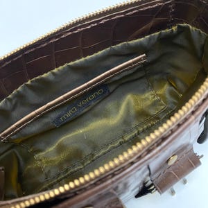 May include: A brown, textured handbag with a gold zipper and hardware. The interior is lined with olive green fabric. The label "mira verano" is visible inside the bag. The bag has a rectangular shape and a crocodile skin pattern.