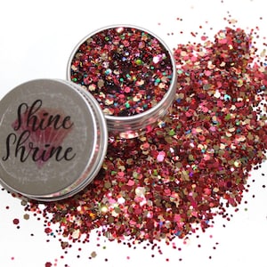 Puede incluir: Dos pequeños botes plateados llenos de purpurina roja, rosa y dorada. En una tapa se lee "Shine Shrine". Parte de la purpurina está esparcida sobre un fondo blanco.