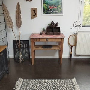Peut inclure: Une table en bois avec un plateau rose et un seul tiroir. La table a une étagère et est décorée d'un vase, de plantes et d'un poster encadré qui dit "Grand Bassin".