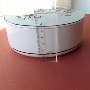 May include: White ceramic round storage container with a black rim and a lid featuring a black and white illustration of a tree with birds and the text "Vaccinium vitis-idaea".
