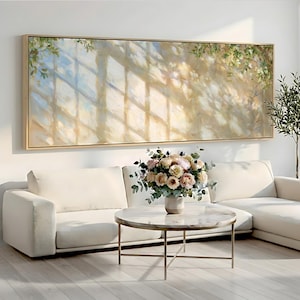 Puede incluir: Un gran cuadro de paisaje rectangular en un marco de madera clara que presenta una representación impresionista y suave de la luz solar filtrándose a través de una ventana sobre el follaje, sobre un sofá seccional blanco y una mesa de centro de mármol.