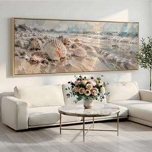 Op de afbeelding: Een grote rechthoekige wandkunstprint met een kustscène met diverse schelpen verspreid over een zandstrand, waarbij zachte golven onder een zacht licht het strand op spoelen, ingelijst in een lichtgekleurde houten lijst.