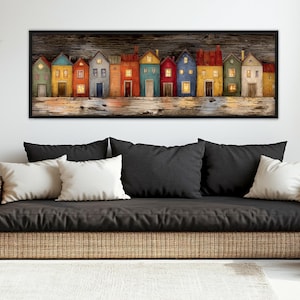 Op de afbeelding: Een ingelijst kunstwerk met een rij kleurrijke huizen met verlichte ramen. De huizen zijn geschilderd in verschillende kleuren, waaronder blauw, geel en rood, tegen een donkere houten achtergrond. Het kunstwerk wordt weergegeven boven een rieten bank met zwarte en witte kussens.