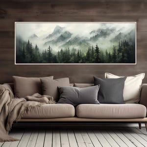 Può includere: Una stampa artistica incorniciata raffigura un paesaggio montano nebbioso con alberi sempreverdi. L'opera d'arte presenta una tavolozza di verdi e grigi, creando un'estetica serena e naturale. La stampa è esposta sopra un divano beige.