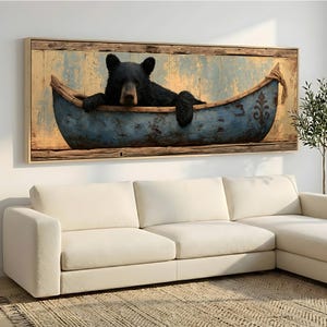Pode incluir: Uma peça de arte de parede retangular emoldurada apresentando um urso preto sentado dentro de uma canoa azul desgastada. A canoa tem um acabamento envelhecido com um emblema de flor-de-lis na lateral. O fundo mostra uma parede de madeira amarela.