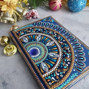 Può includere: Un quaderno blu con un disegno di mandala dipinto sulla copertina. Il disegno presenta accenti dorati, bianchi e turchesi. Il quaderno è circondato da decorazioni natalizie dorate e blu.