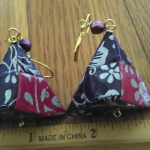 May include: A pair of handmade earrings with a triangular shape. The earrings are made of fabric with a floral pattern in shades of purple, red, and white. The earrings are accented with gold wire and purple beads.