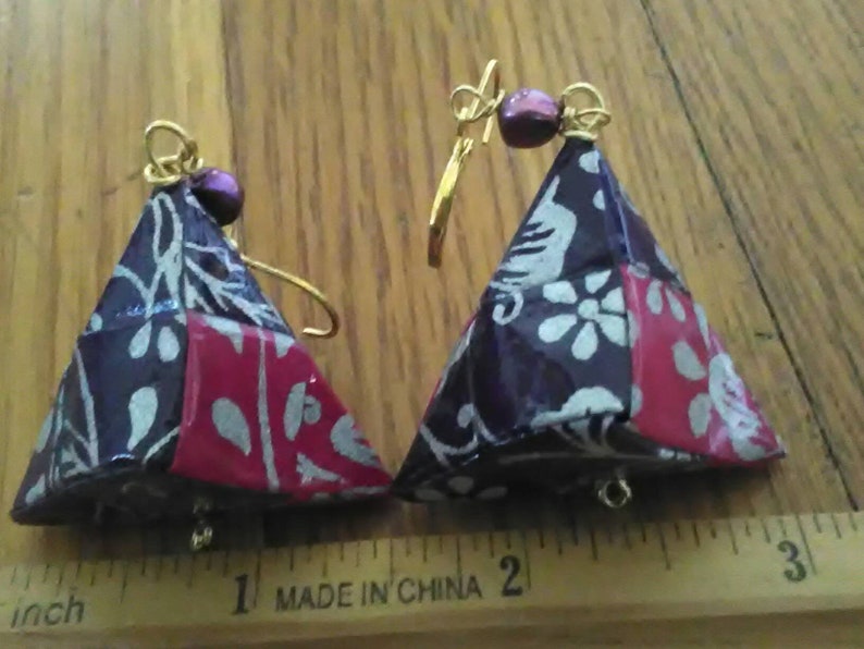 May include: A pair of handmade earrings with a triangular shape. The earrings are made of fabric with a floral pattern in shades of purple, red, and white. The earrings are accented with gold wire and purple beads.