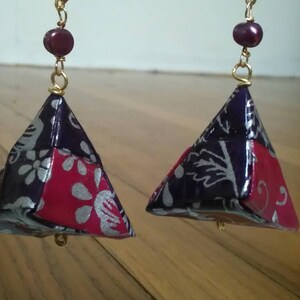 May include: A pair of purple and pink origami earrings with a silver floral pattern. The earrings are suspended from gold chains with a single purple bead at the top of each chain.