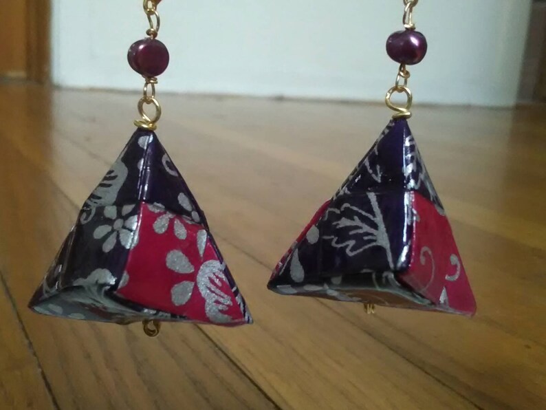 May include: A pair of purple and pink origami earrings with a silver floral pattern. The earrings are suspended from gold chains with a single purple bead at the top of each chain.