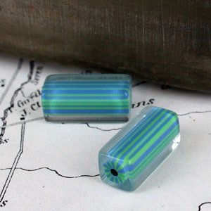May include: Two clear glass beads with blue and green stripes. The beads are rectangular in shape and have a translucent appearance.
