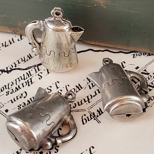 May include: Three small, silver-colored teapot charms. Each charm has a handle, spout, and a decorative wavy line around the middle. The charms are displayed on a vintage map.