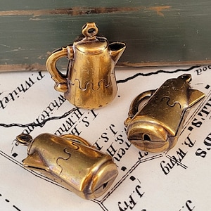 May include: Three small, antique-style brass teapots. Each teapot has a handle, spout, and a decorative wavy line near the base. The teapots are arranged on a vintage map background.