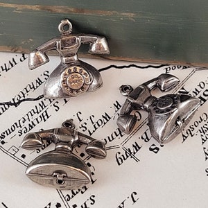 Puede incluir: Tres colgantes de teléfono de metal plateado. Cada colgante tiene un dial giratorio y un auricular. Los colgantes están detallados con una estética vintage. El fondo incluye un mapa y una superficie verde.