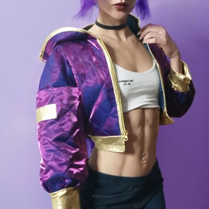 Può includere: Una giacca corta viola e oro con un design trapuntato e una chiusura a zip. La giacca ha un cappuccio e una toppa sulla manica sinistra. Il testo "Los Angeles" è visibile sul retro della giacca.