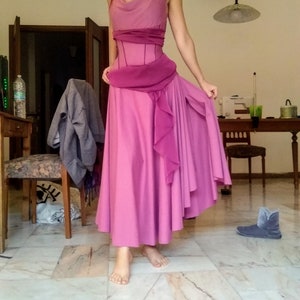May include: A floor-length sleeveless dress in shades of pink and magenta, featuring a cowl neckline, a fitted bodice with vertical seams, and a draped sash at the waist. The skirt is full and flowing with a ruffled side panel and a high slit.