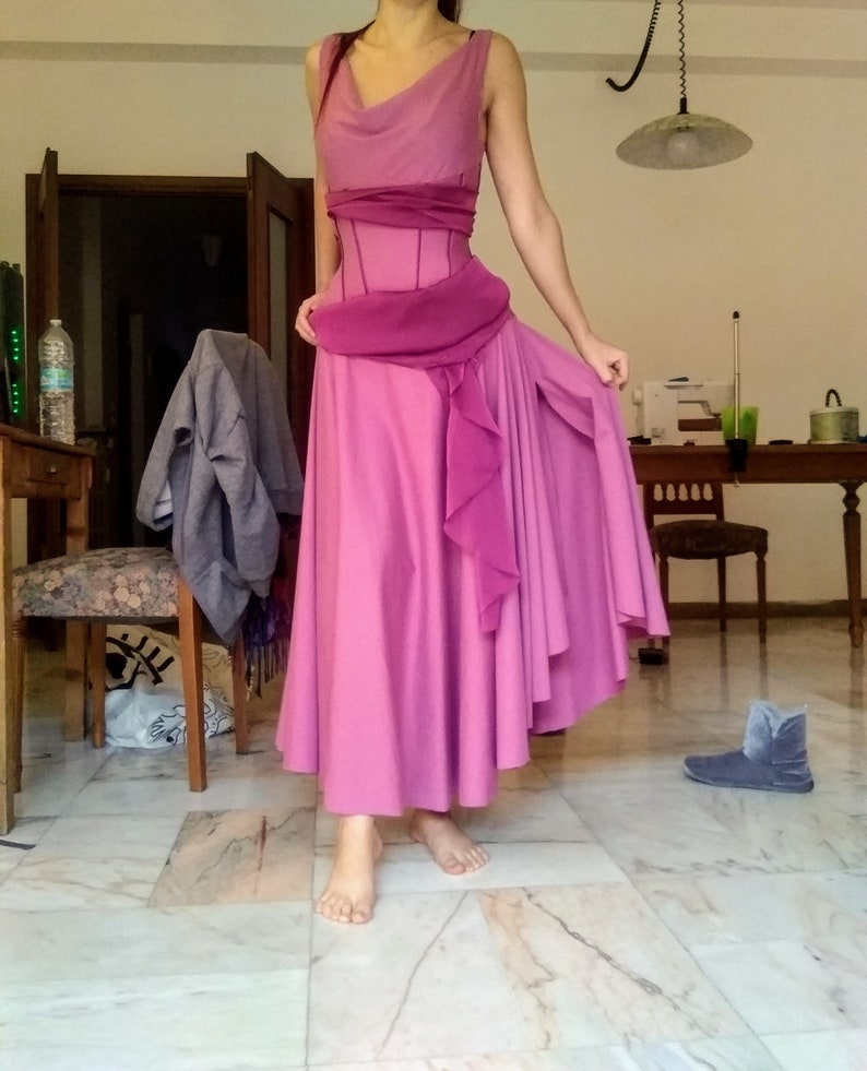May include: A floor-length sleeveless dress in shades of pink and magenta, featuring a cowl neckline, a fitted bodice with vertical seams, and a draped sash at the waist. The skirt is full and flowing with a ruffled side panel and a high slit.