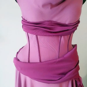 May include: A mauve dress featuring a structured corset-style bodice with vertical boning channels. The garment includes a draped cowl neckline and a wide, gathered fabric sash wrapped horizontally around the waist, creating a layered effect.
